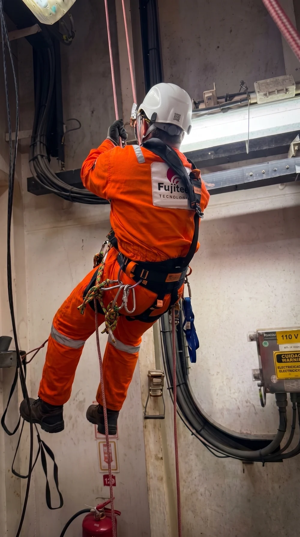 Fujitec technician in orange coveralls working at height on rope access, next to cable trays and an electrical box